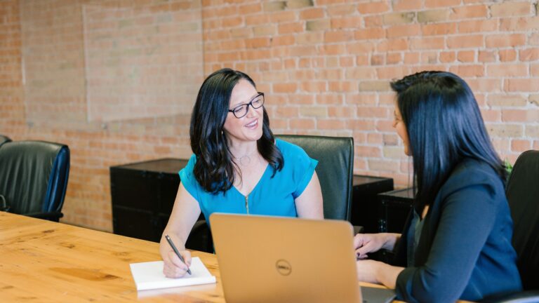 Two businesswomen talking one with notepad and the other with an open laptop