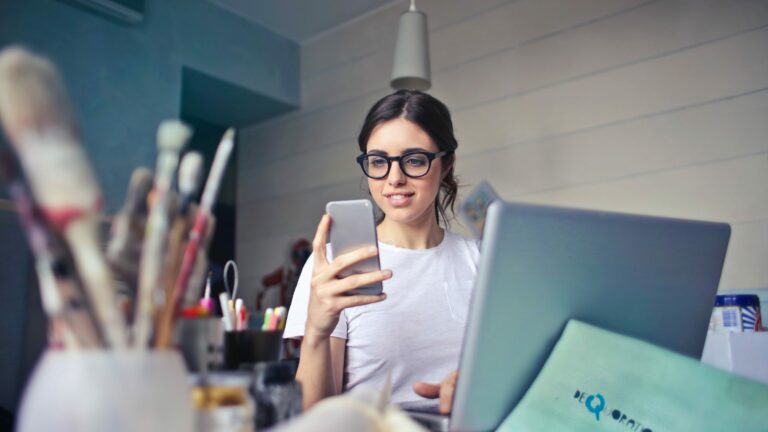 young women wearing spectacles looking at mobile phone while sitting at desk with laptop