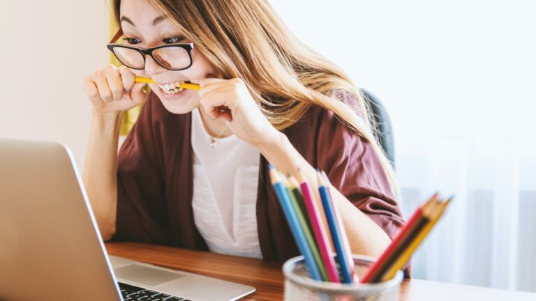 young woman wearing spectacle, staring at laptop whilst holding a pencil in her mouth