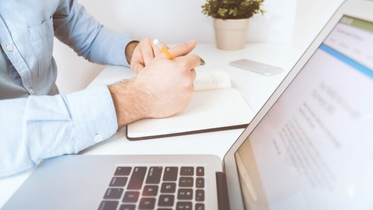 man writing in notebook with laptop in foreground