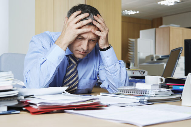 businessman with head in hands sitting at desk filled with documents and papers