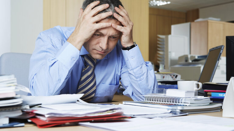 businessman with head in hands sitting at desk filled with documents and papers