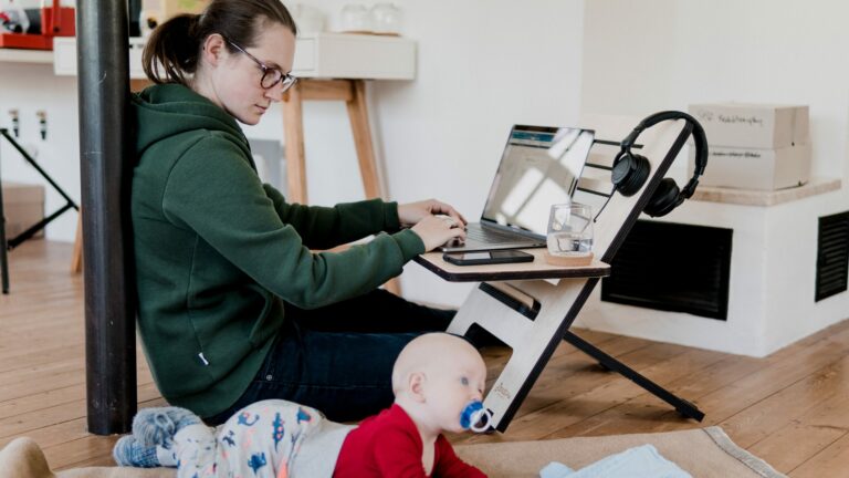 woman sitting on floor, typing on laptop with baby on floor beside her