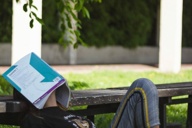 young person laying on grass outside with head resting on bench and face covered with an open book