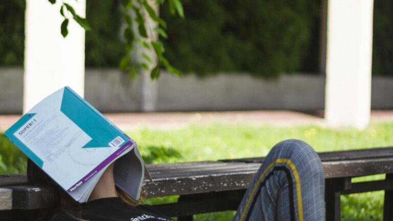 young person laying on grass outside with head resting on bench and face covered with an open book