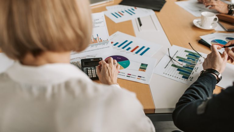 Woman seen from behind using a calculator on desk covered with diagrams of graphs.